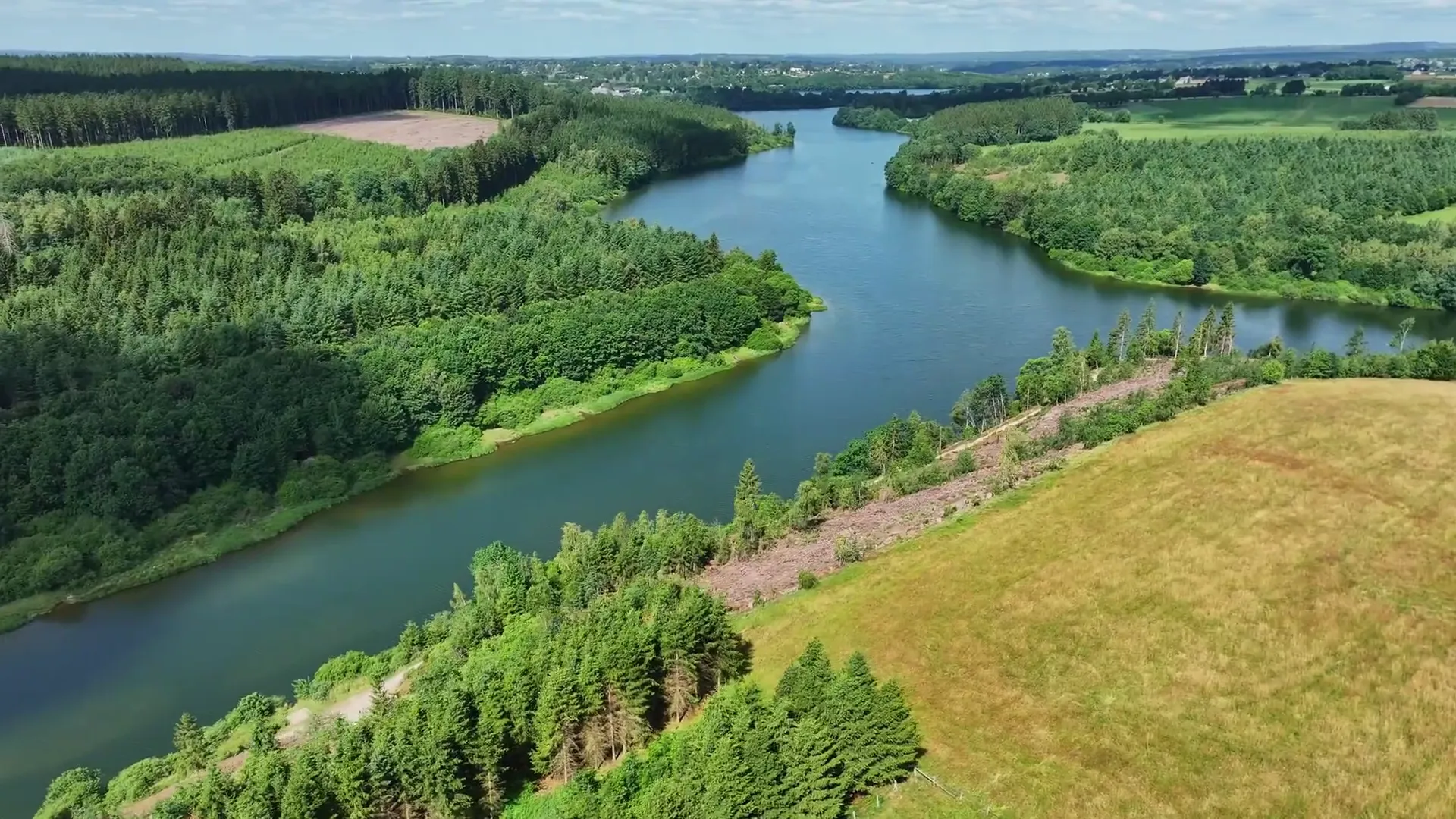 Vue aérienne d'un lac serpentant entre des collines boisées, une prairie au premier plan
