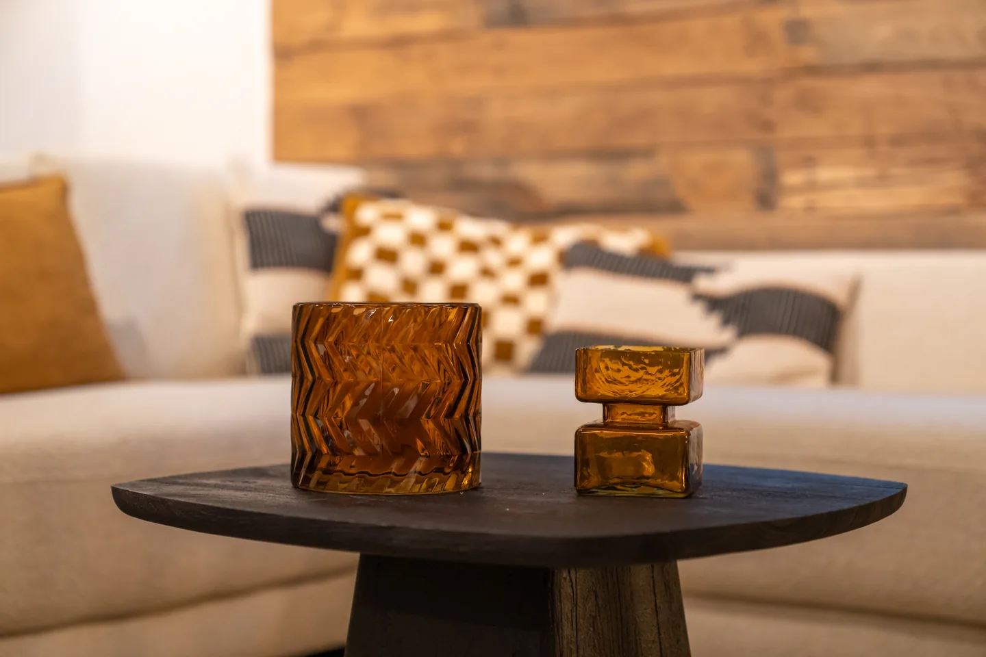 Coin salon chaleureux : canapé beige, coussins à motifs moutarde et crème, mur en bois brut, décor en verre ambré