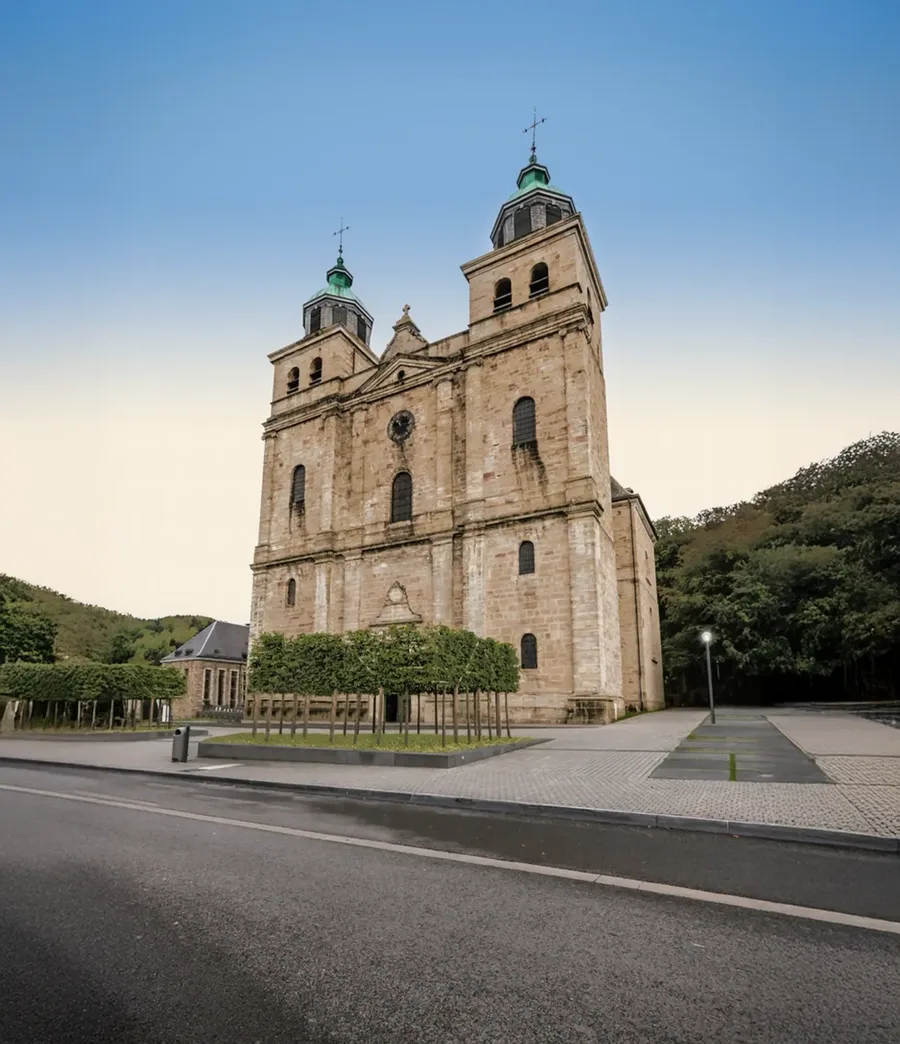 Die Kathedrale von Malmedy mit ihren zwei Türmen vor einem bewaldeten Hügel