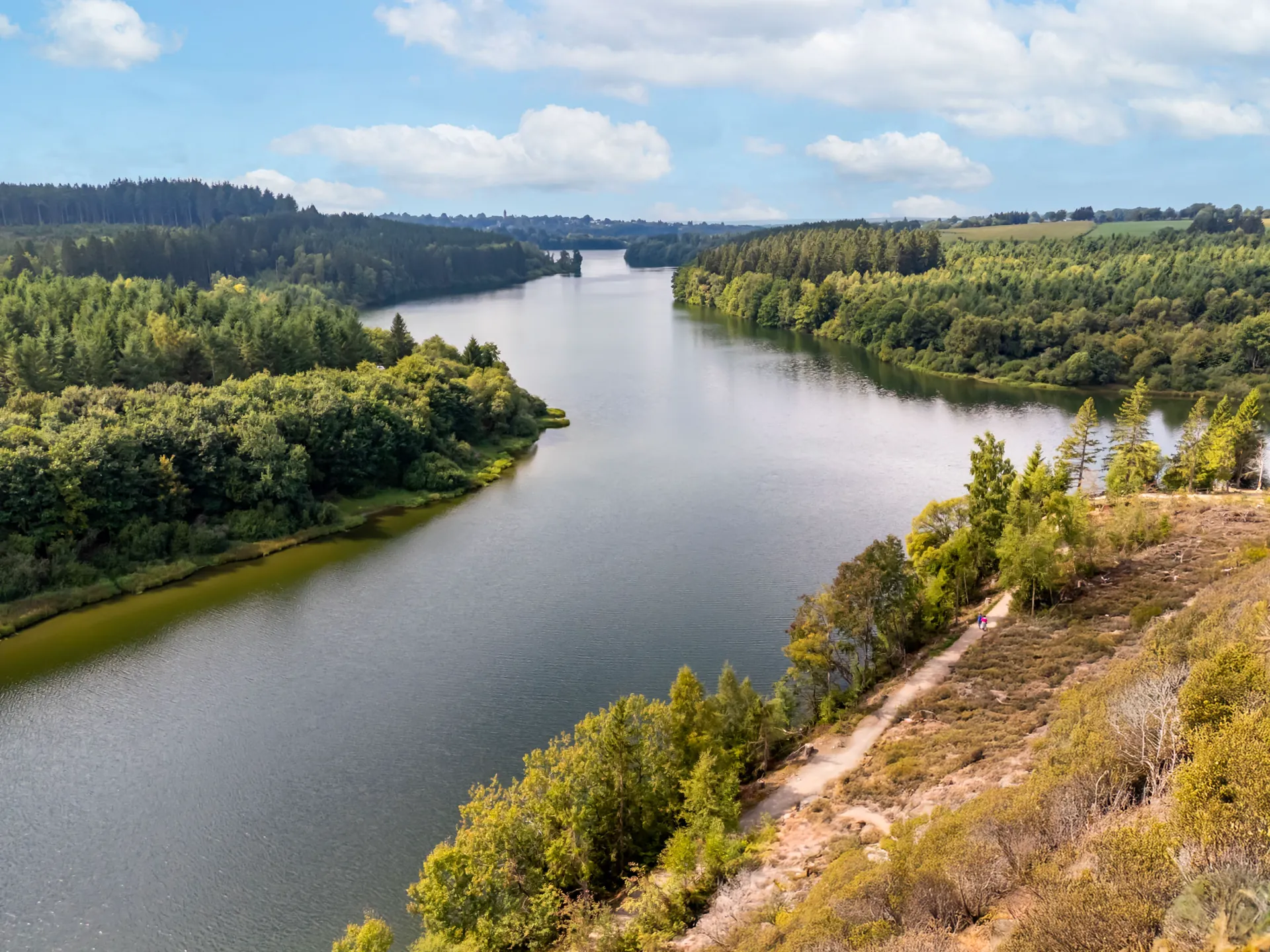 Vue aérienne du lac serpentant entre des collines boisées, un sentier longeant la rive