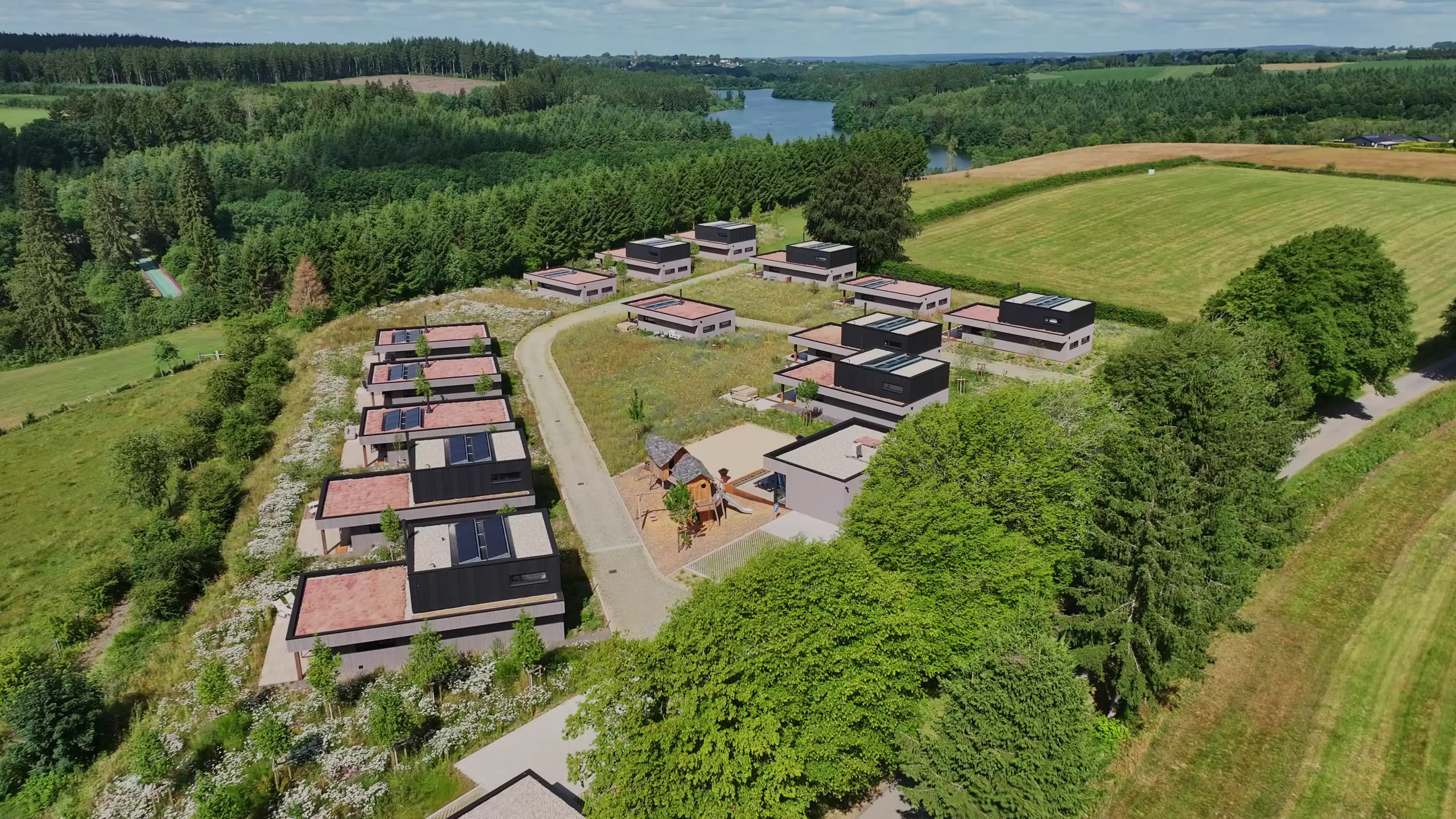 Vue aérienne du domaine Nature Lake Village : les villas alignées le long du chemin, la Welcome villa et l’aire de jeux au centre, le parking et la forêt en arrière-plan.