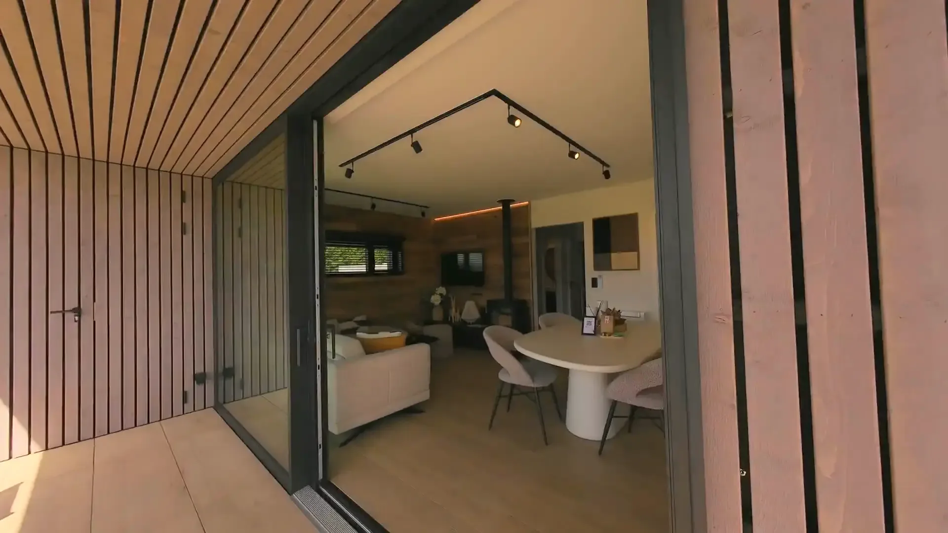 View from the covered wooden terrace through the open sliding door into the living and dining area with round table, sofa and wood stove