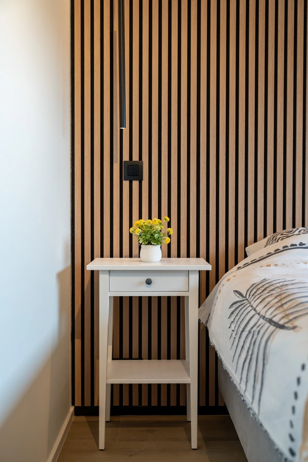 Schlafzimmer: Nachttisch mit Blumenstrauß vor einer Wand aus vertikalen Holzlatten, Bett im Hintergrund