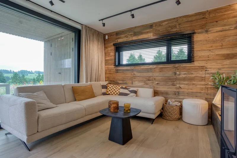 Wohnzimmer mit Ecksofa, Holzofen und großer Fensterfront zur Wiese