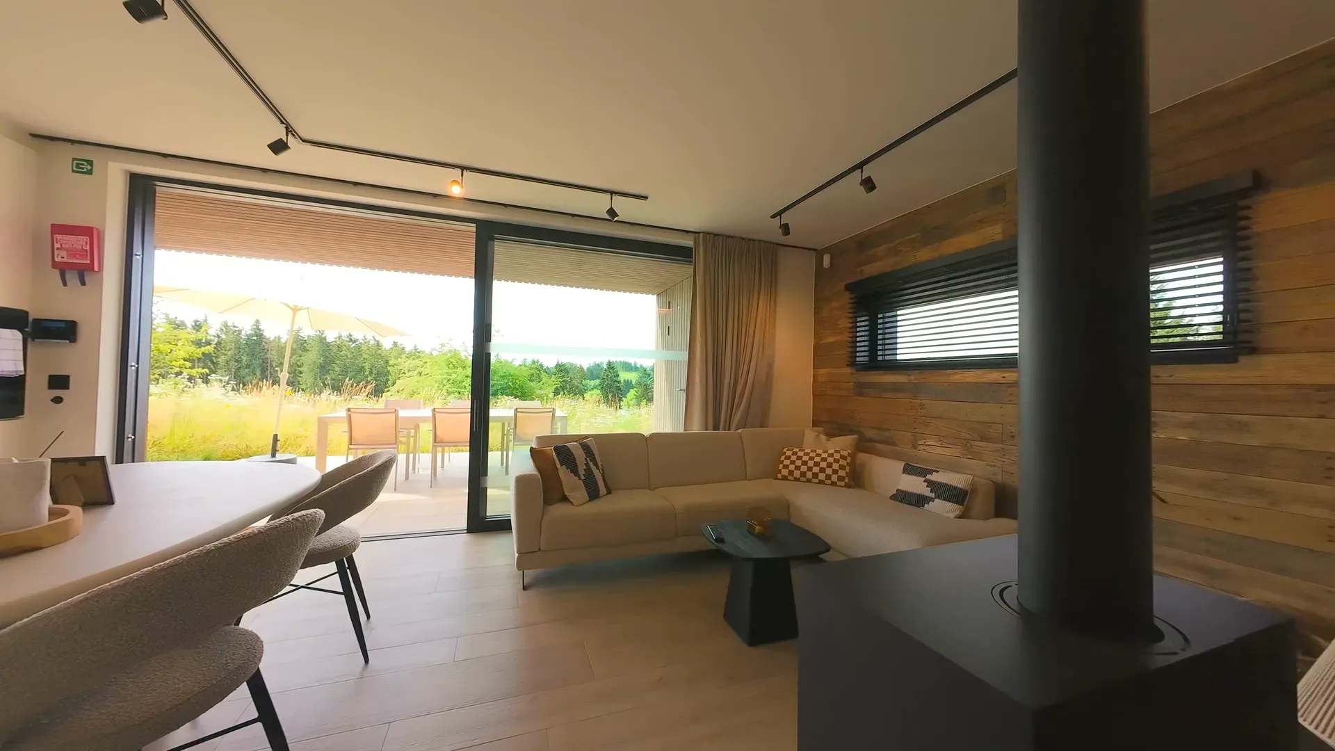 Séjour avec canapé d'angle et poêle à bois, vue à travers la baie coulissante ouverte sur la terrasse et sa table dressée à la lumière du soir