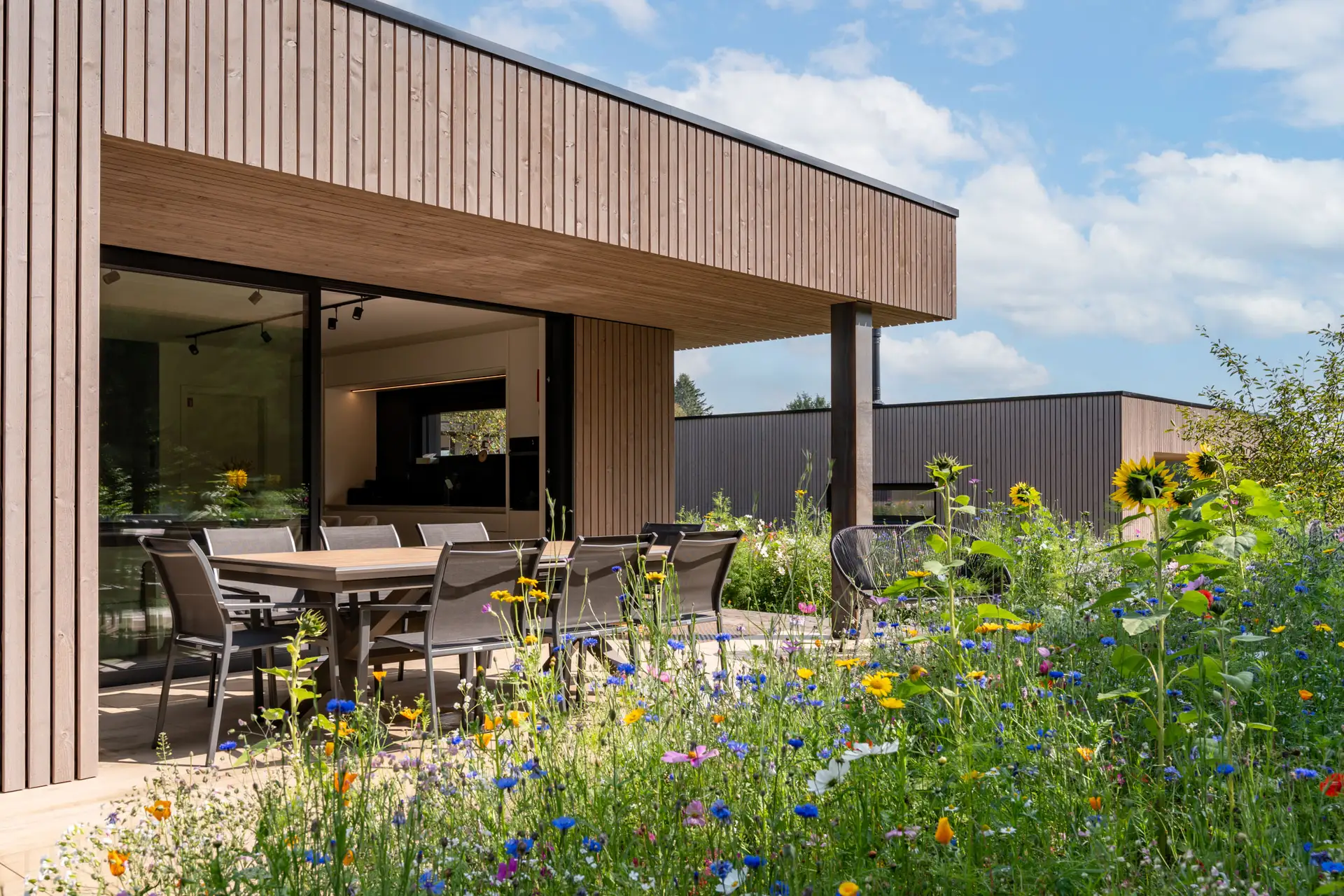 Terrasse couverte avec grande table à manger, prairie fleurie devant la villa